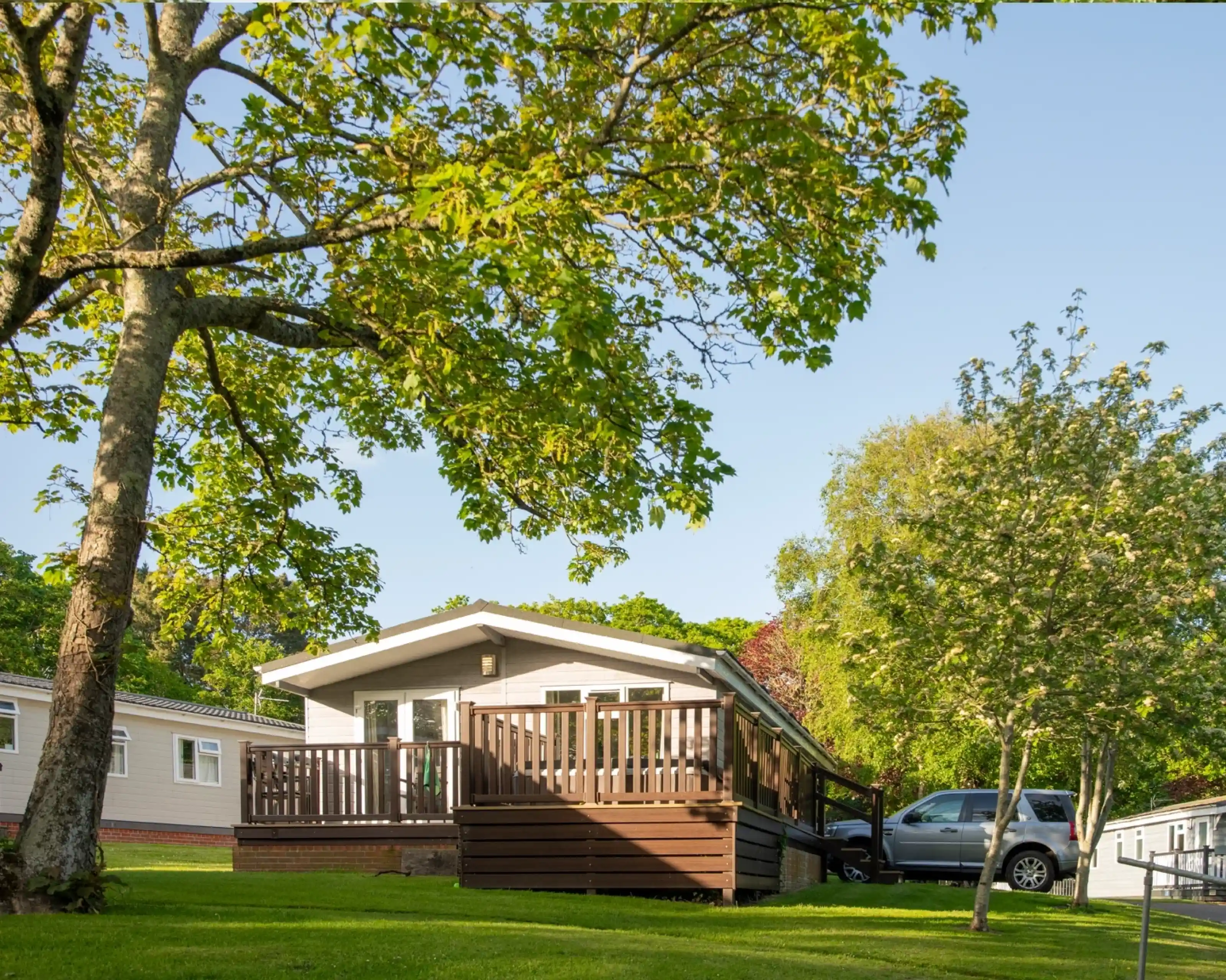 A modern chalet-style home with a wooden deck surrounded by green trees and a well-manicured lawn. A car is parked nearby, and additional mobile homes can be seen in the background under clear blue skies.