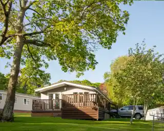 A modern chalet-style home with a wooden deck surrounded by green trees and a well-manicured lawn. A car is parked nearby, and additional mobile homes can be seen in the background under clear blue skies.