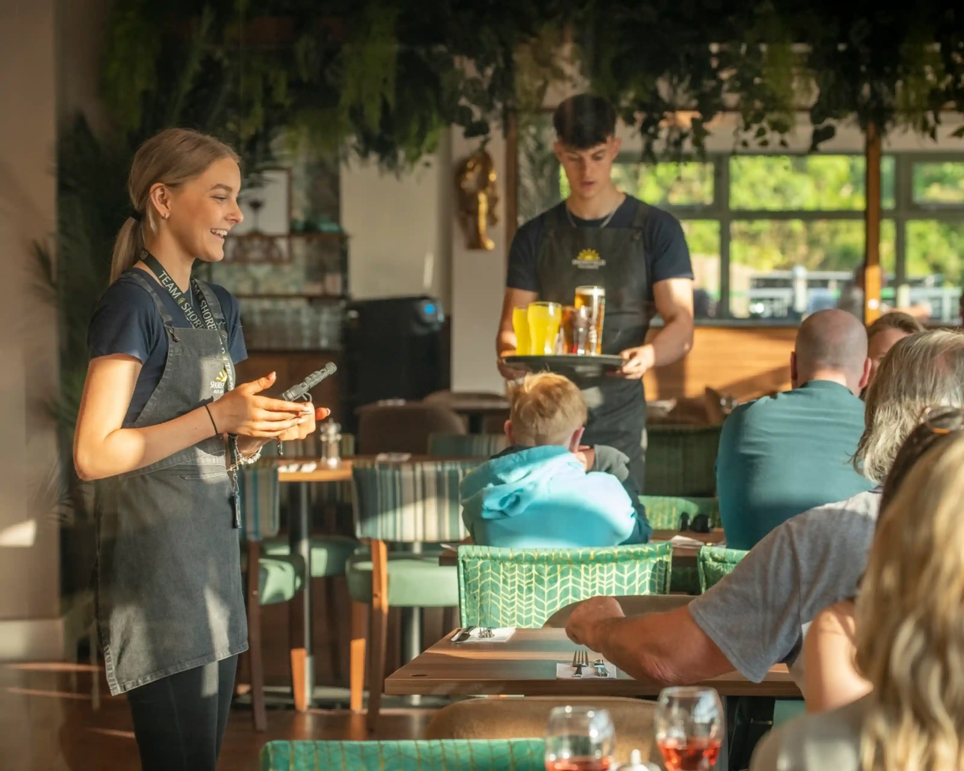A young female server stands smiling, holding a tablet, while a male server carries a tray with drinks. Diners are seated at tables, with a light-filled restaurant atmosphere featuring greenery in the background.