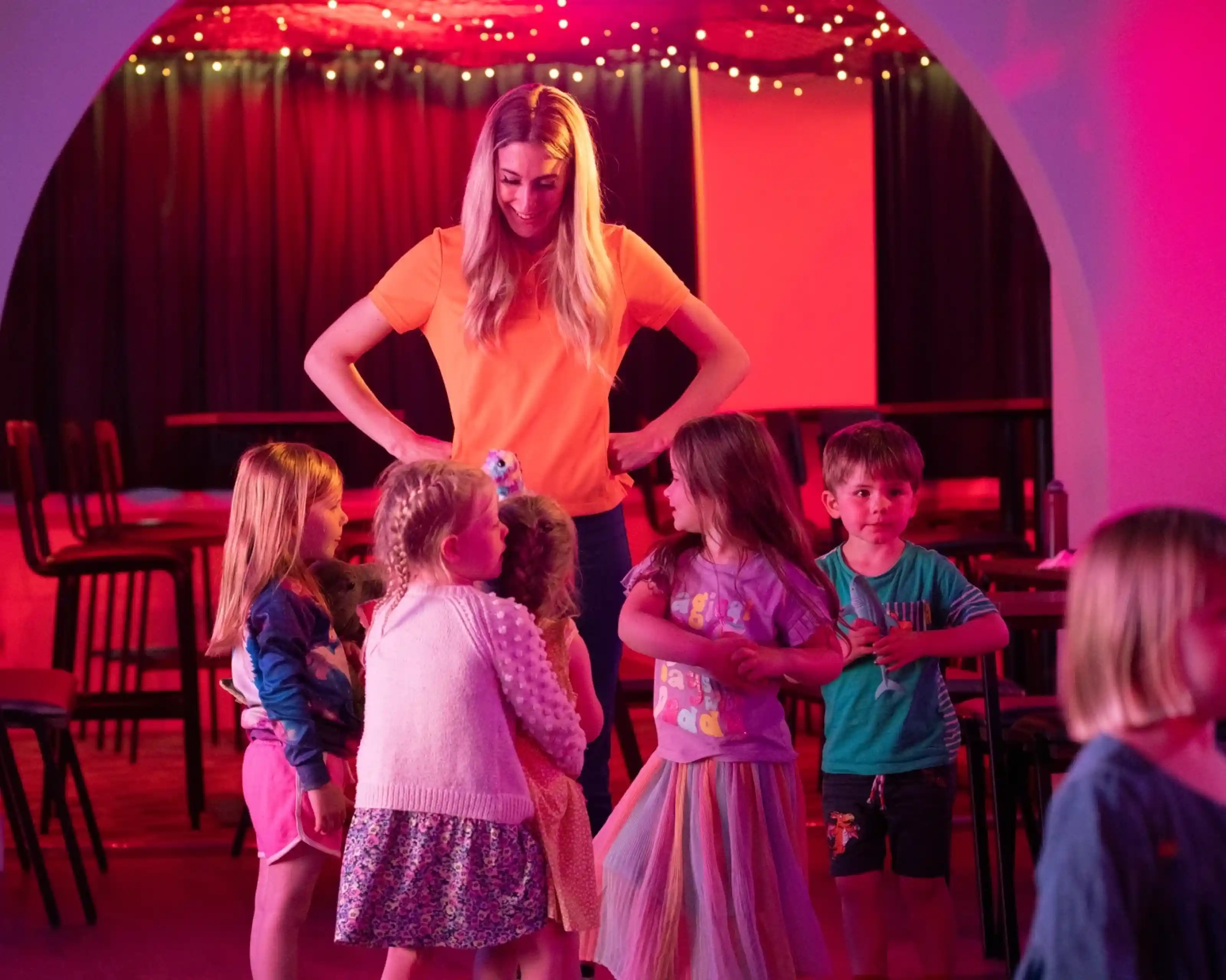 A group of children gather around a woman with long blonde hair, who stands confidently with her hands on her hips. The setting features colorful lights and a cozy atmosphere, with several chairs in the background and the children engaged and smiling.