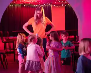 A group of children gather around a woman with long blonde hair, who stands confidently with her hands on her hips. The setting features colorful lights and a cozy atmosphere, with several chairs in the background and the children engaged and smiling.