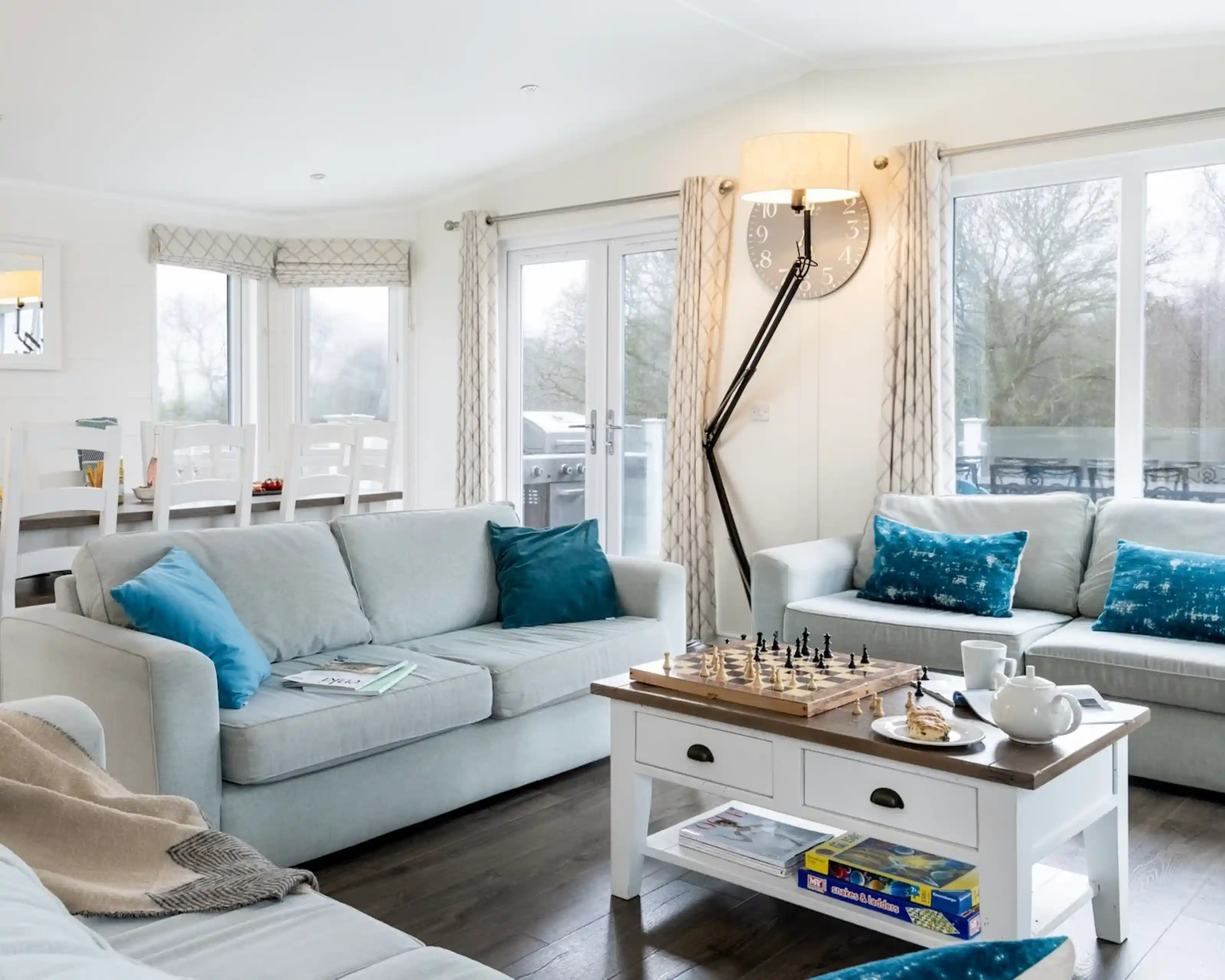 A bright, modern living room featuring two light gray sofas adorned with blue pillows. A wooden coffee table with a chess set sits in the center. Soft, natural light streams through large windows, revealing a view of greenery outside. A dining area with white chairs is visible in the background.