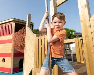 A young boy in an orange shirt smiles confidently while climbing a shiny pole on a colorful playground. Bright blue sky and green trees are in the background.