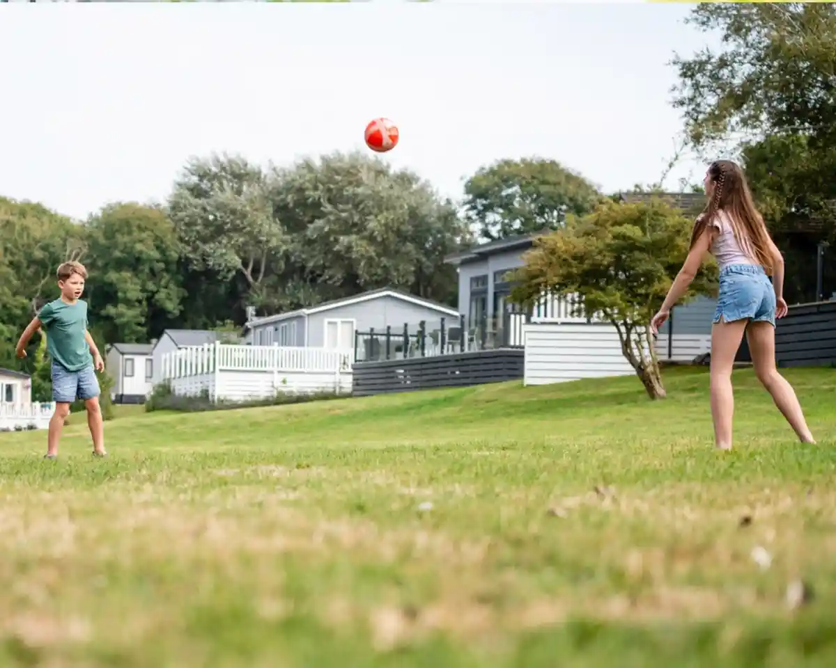 Two children playing catch with a red ball on a grassy area. They are smiling, with houses and trees in the background. The boy wears a green shirt and the girl wears a tank top and shorts.