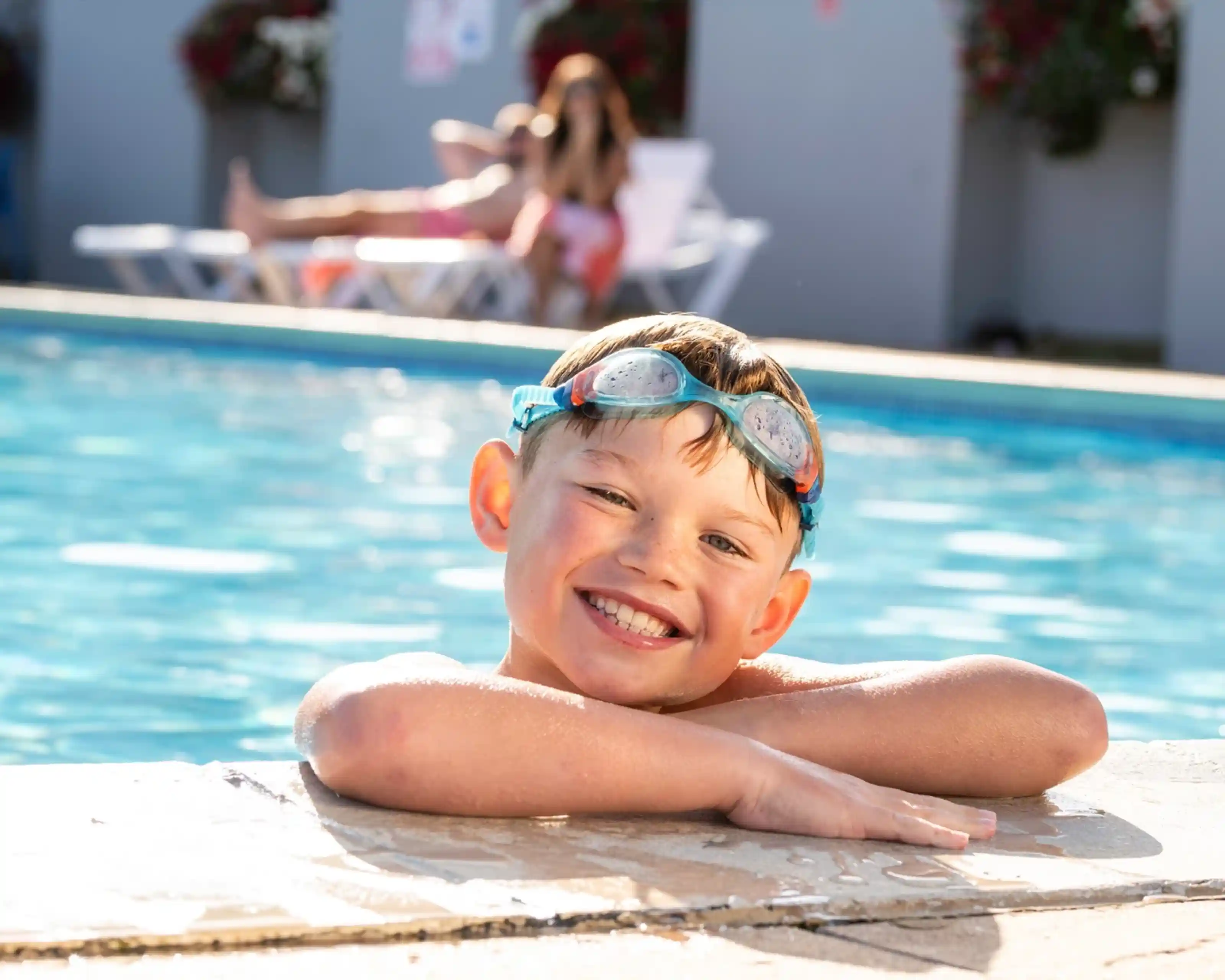 A smiling boy with swim goggles leans on the edge of a swimming pool. In the background, a person relaxes on a lounge chair. Colorful flowers adorn the walls nearby.