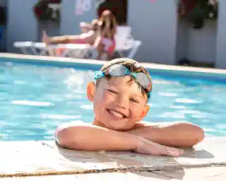 A smiling boy with swim goggles leans on the edge of a swimming pool. In the background, a person relaxes on a lounge chair. Colorful flowers adorn the walls nearby.