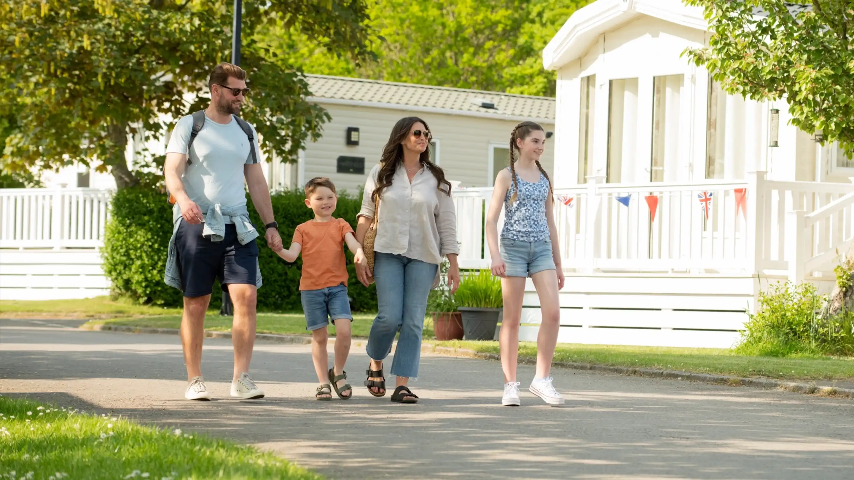 A family of four walks together along a tree-lined pathway. The father, wearing a backpack, holds hands with his young son, while the mother walks beside them, holding hands with their teenage daughter. They all appear relaxed and happy, enjoying a sunny day. In the background, there are white trailers and greenery.