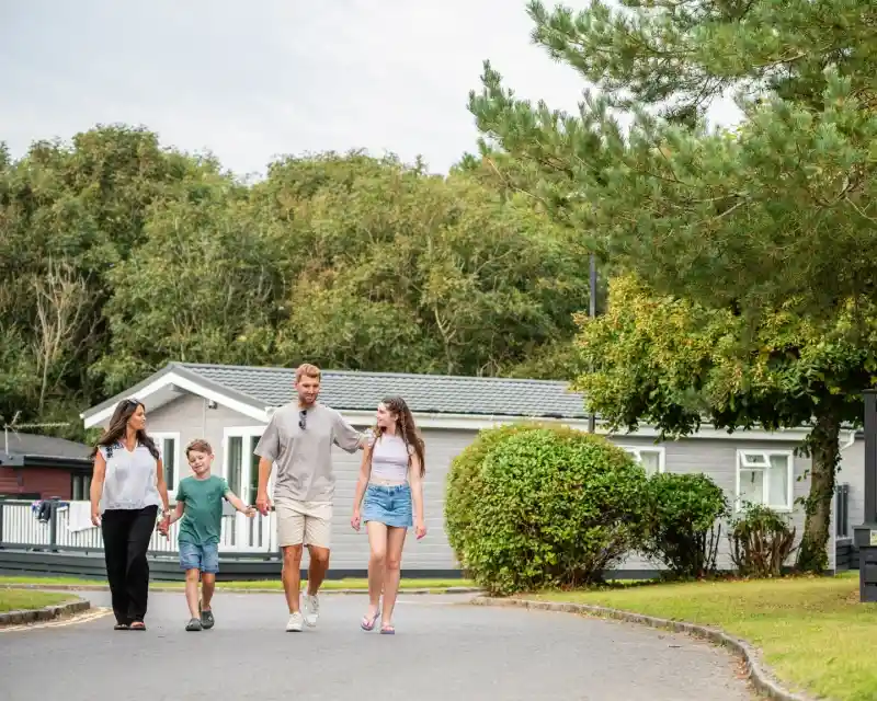 A family of four walks together along a path in a residential area. A woman and a young boy hold hands, while a man walks with a teenage girl. Lush greenery and homes are visible in the background.