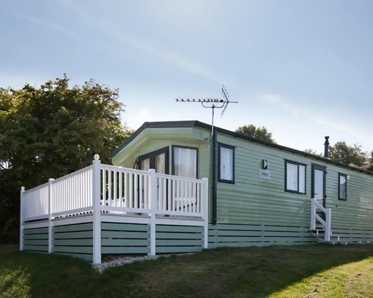 A light green mobile home with a white railing surrounding a deck, sitting on a grassy area. There is an antenna on the roof and trees in the background under a clear blue sky.