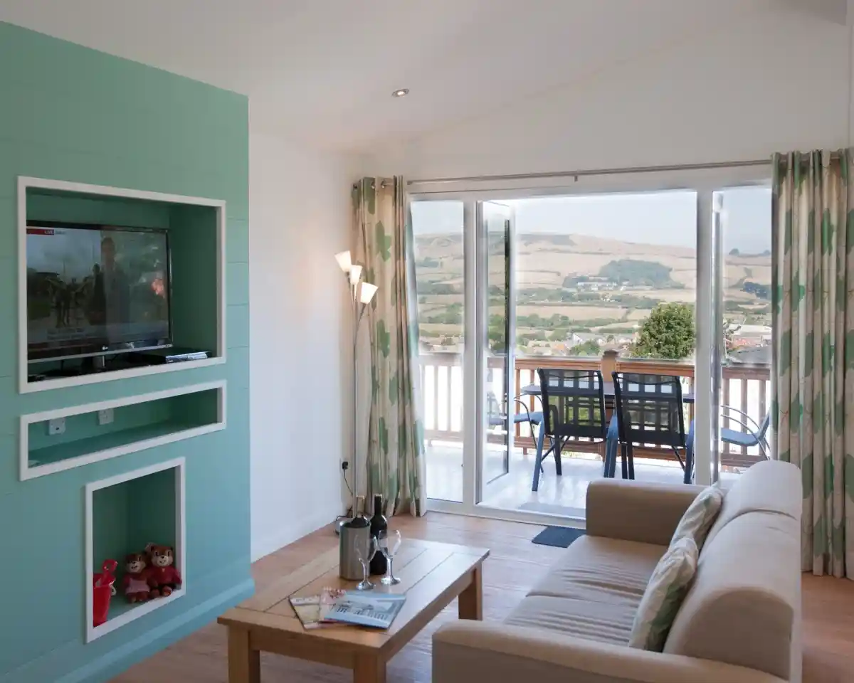 A cozy living room with a light green accent wall featuring a mounted television. A beige sofa faces a coffee table with a magazine and two bottles. Glass doors open to a balcony with outdoor seating and scenic views of hills and trees.