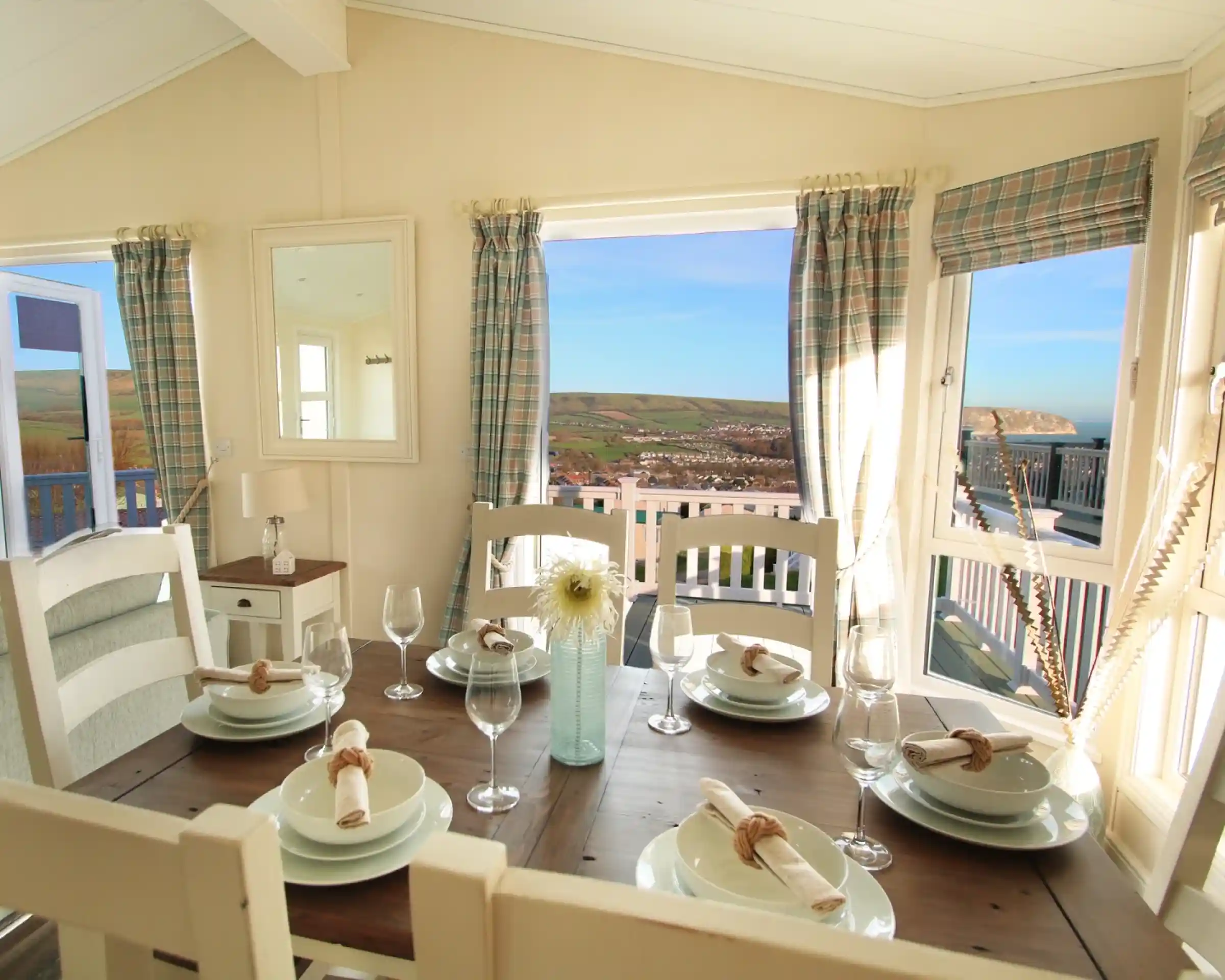 A bright dining area with a wooden table set for six, featuring white plates, glassware, and rolled napkins. Large windows offer a view of hills and a blue sky, while plaid curtains frame the scene. A mirror and a vase with a flower decorate the space.