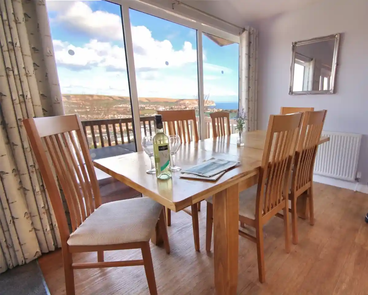 A spacious dining table made of wood is set with six chairs, positioned by a large window showcasing a scenic view of a coastal landscape. A bottle of wine sits on the table, and a decorative mirror is visible on the wall. Natural light floods the room, complementing the wood floors and neutral decor.