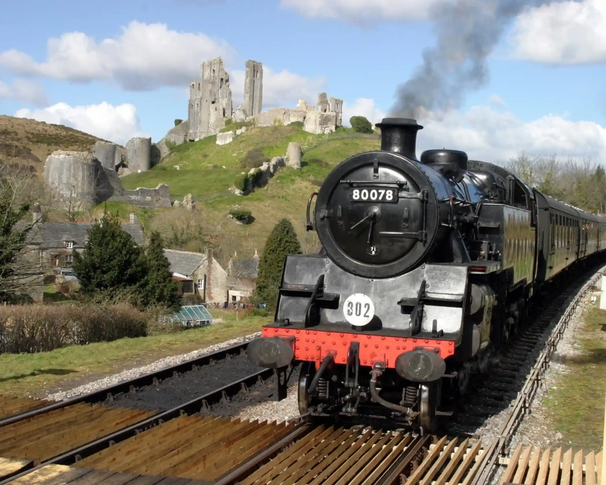 A vintage steam locomotive travels along tracks in the foreground, with a backdrop of a historic castle and rolling hills under a partly cloudy sky.