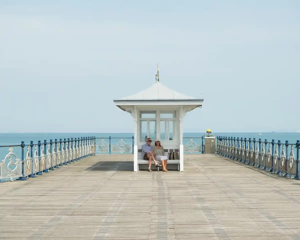 A couple sits together on a wooden pier near a small white gazebo, with a calm sea visible in the background. The sky is clear and blue, creating a serene atmosphere.