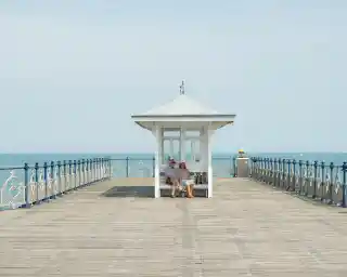 A couple sits together on a wooden pier near a small white gazebo, with a calm sea visible in the background. The sky is clear and blue, creating a serene atmosphere.