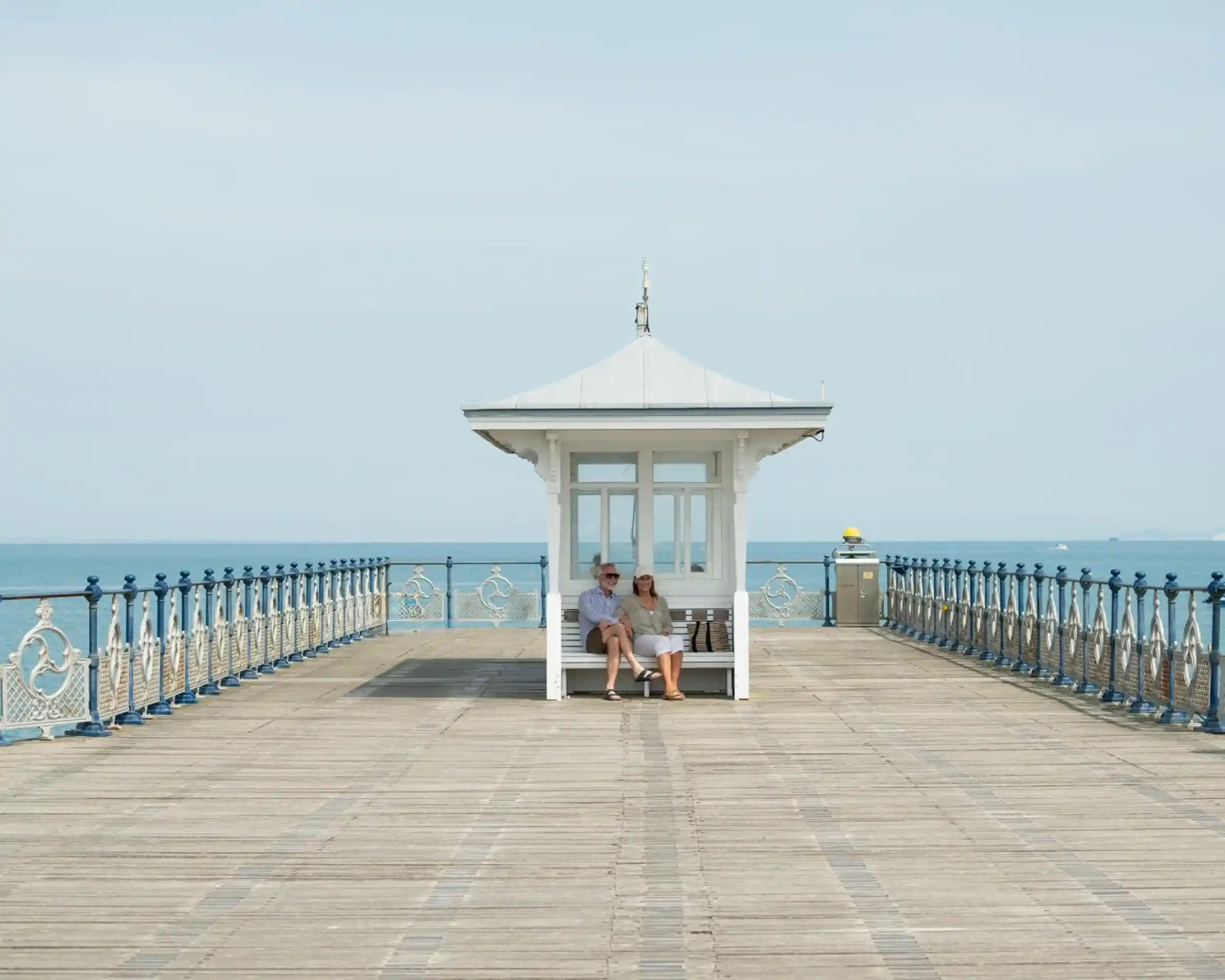 A couple sits together on a wooden pier near a small white gazebo, with a calm sea visible in the background. The sky is clear and blue, creating a serene atmosphere.