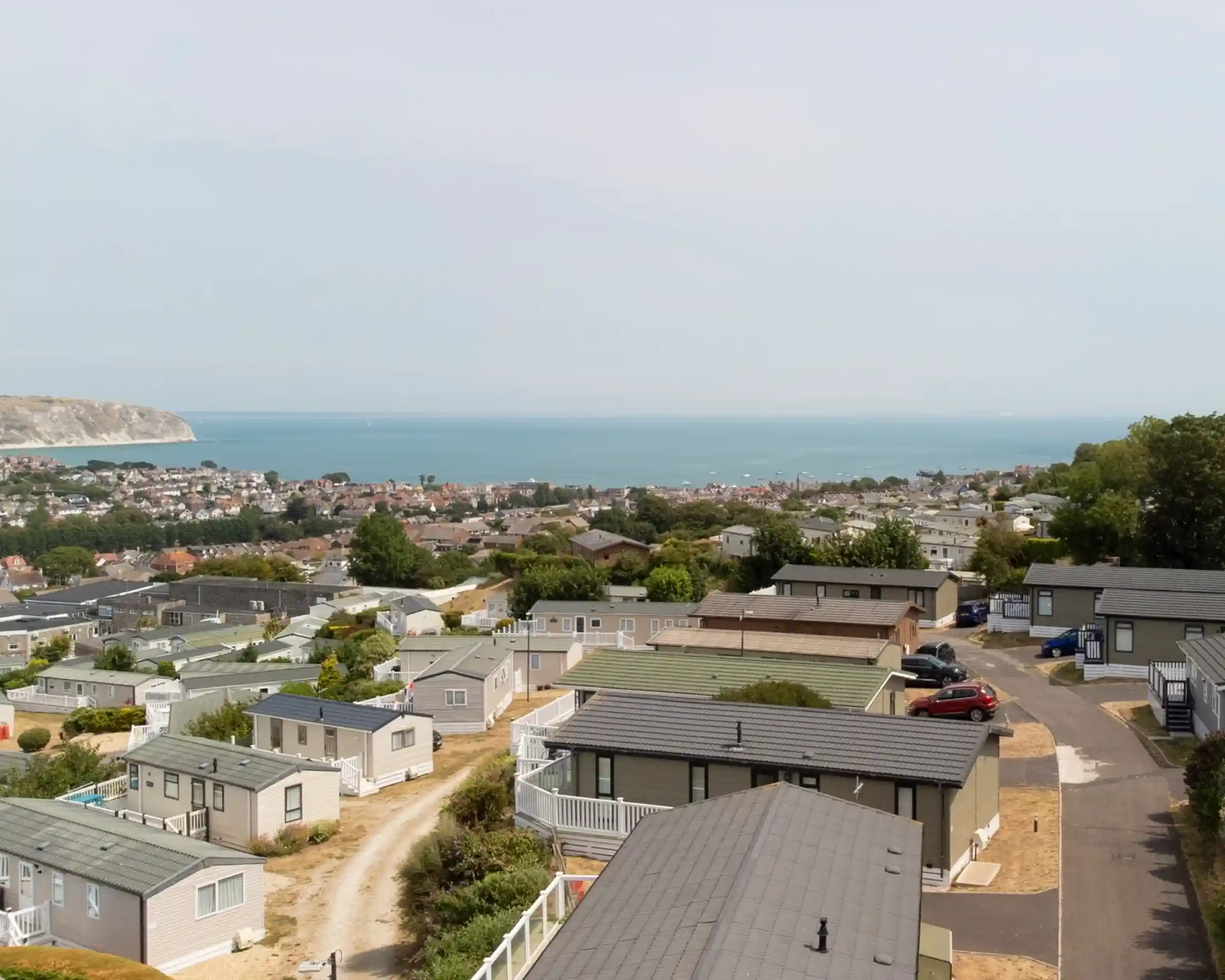 A panoramic view of a coastal area featuring rows of holiday homes with ocean and distant hills in the background. The sky is slightly overcast, and a few cars are parked along the winding road.