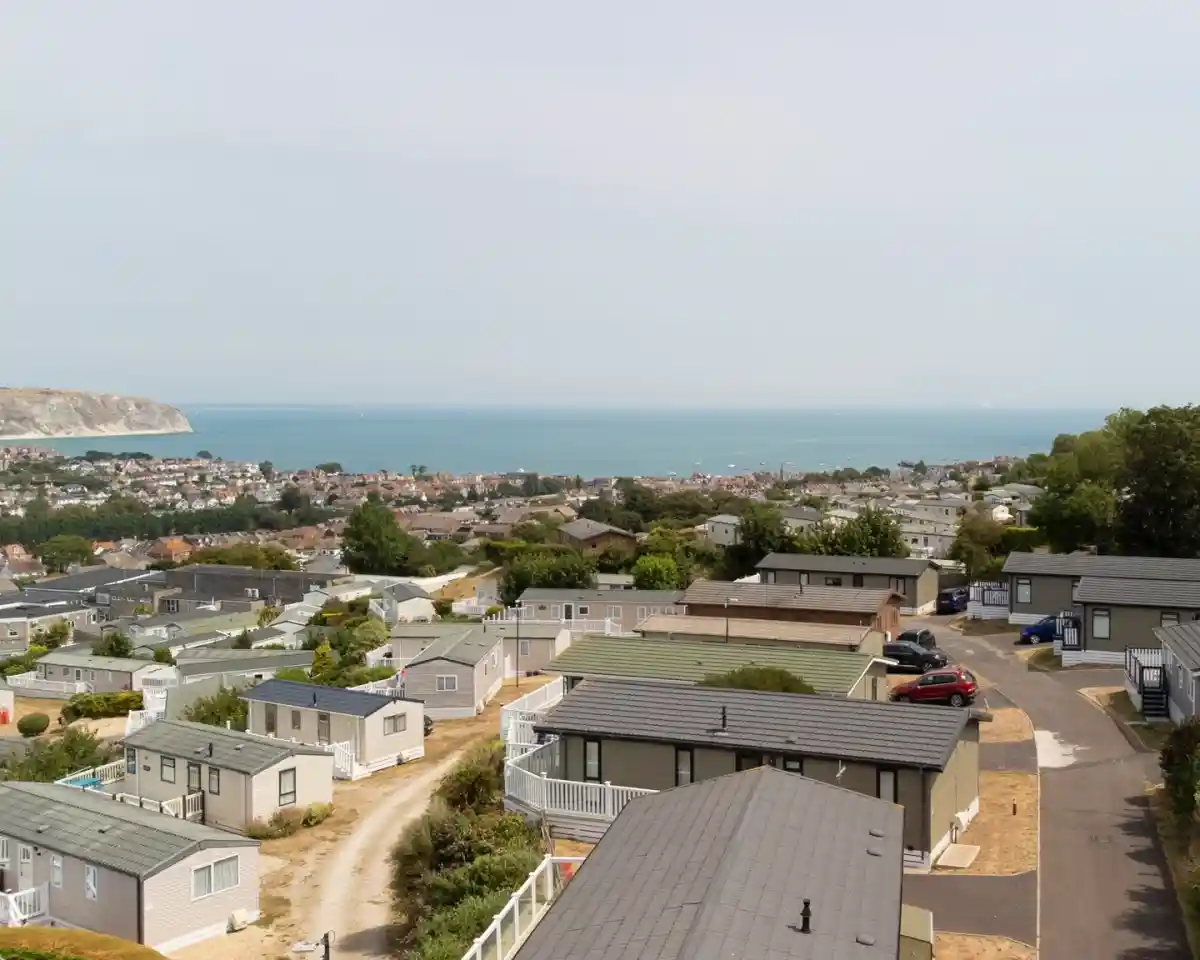 A panoramic view of a coastal area featuring rows of holiday homes with ocean and distant hills in the background. The sky is slightly overcast, and a few cars are parked along the winding road.