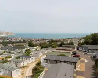 A panoramic view of a coastal area featuring rows of holiday homes with ocean and distant hills in the background. The sky is slightly overcast, and a few cars are parked along the winding road.