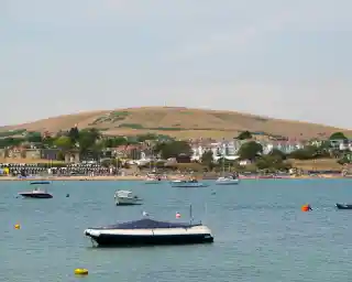 A tranquil seaside scene featuring several boats floating in calm water, with a sandy beach and residential buildings in the background. A large green hill rises in the distance under a clear sky.