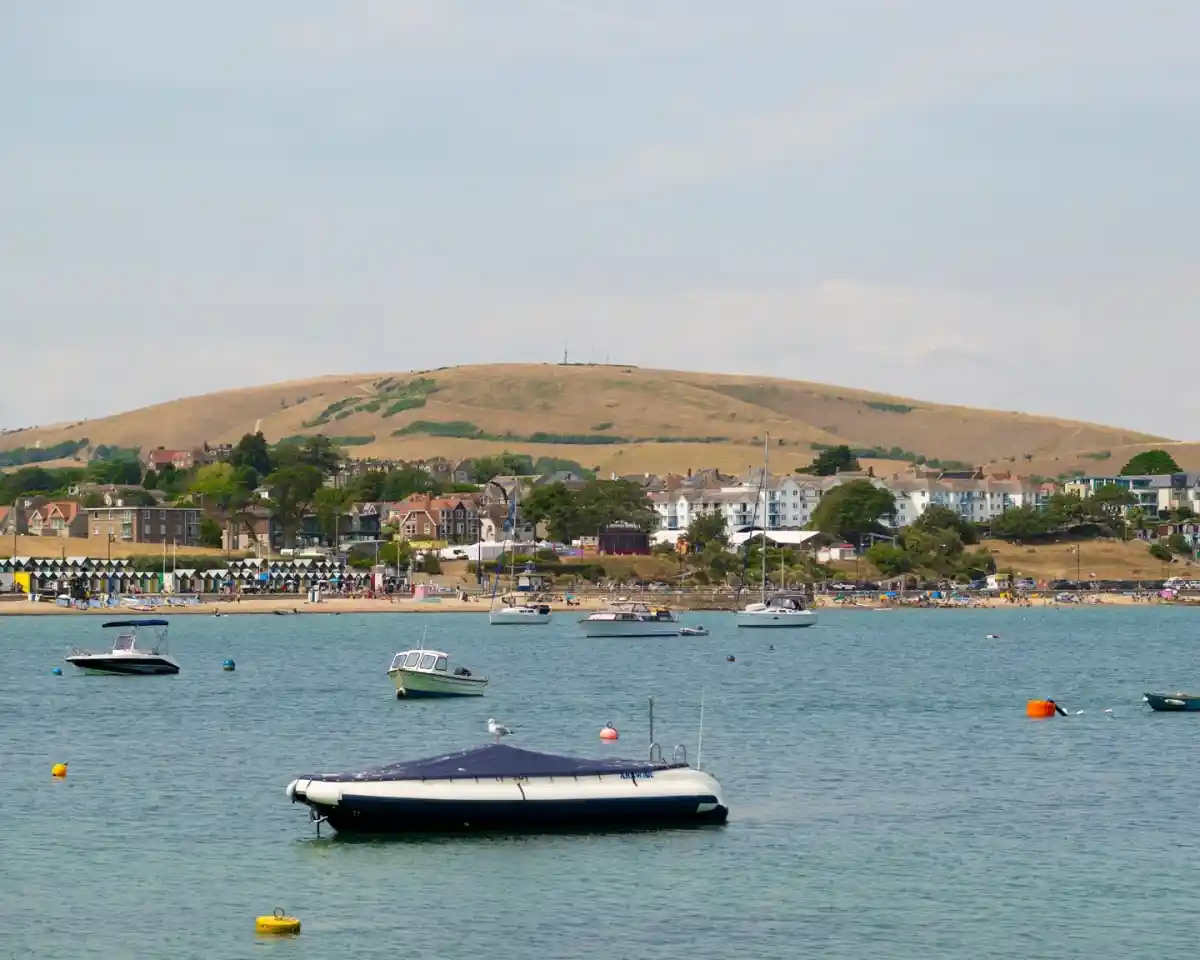 A tranquil seaside scene featuring several boats floating in calm water, with a sandy beach and residential buildings in the background. A large green hill rises in the distance under a clear sky.