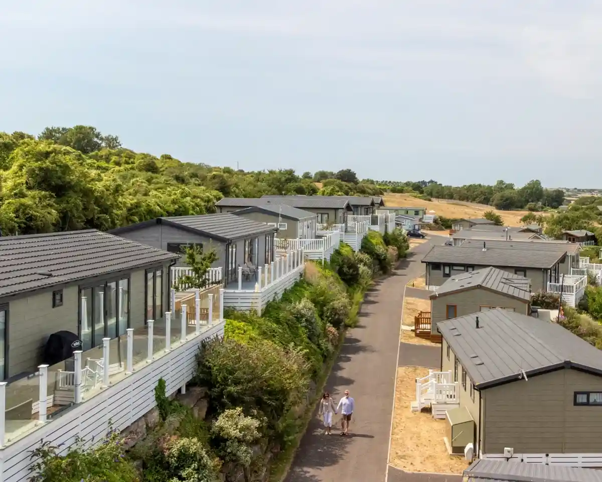 Aerial view of a residential area featuring several modern holiday lodges along a winding road. Two people walk hand-in-hand along the pathway. Lush greenery surrounds the lodges, providing a serene atmosphere.
