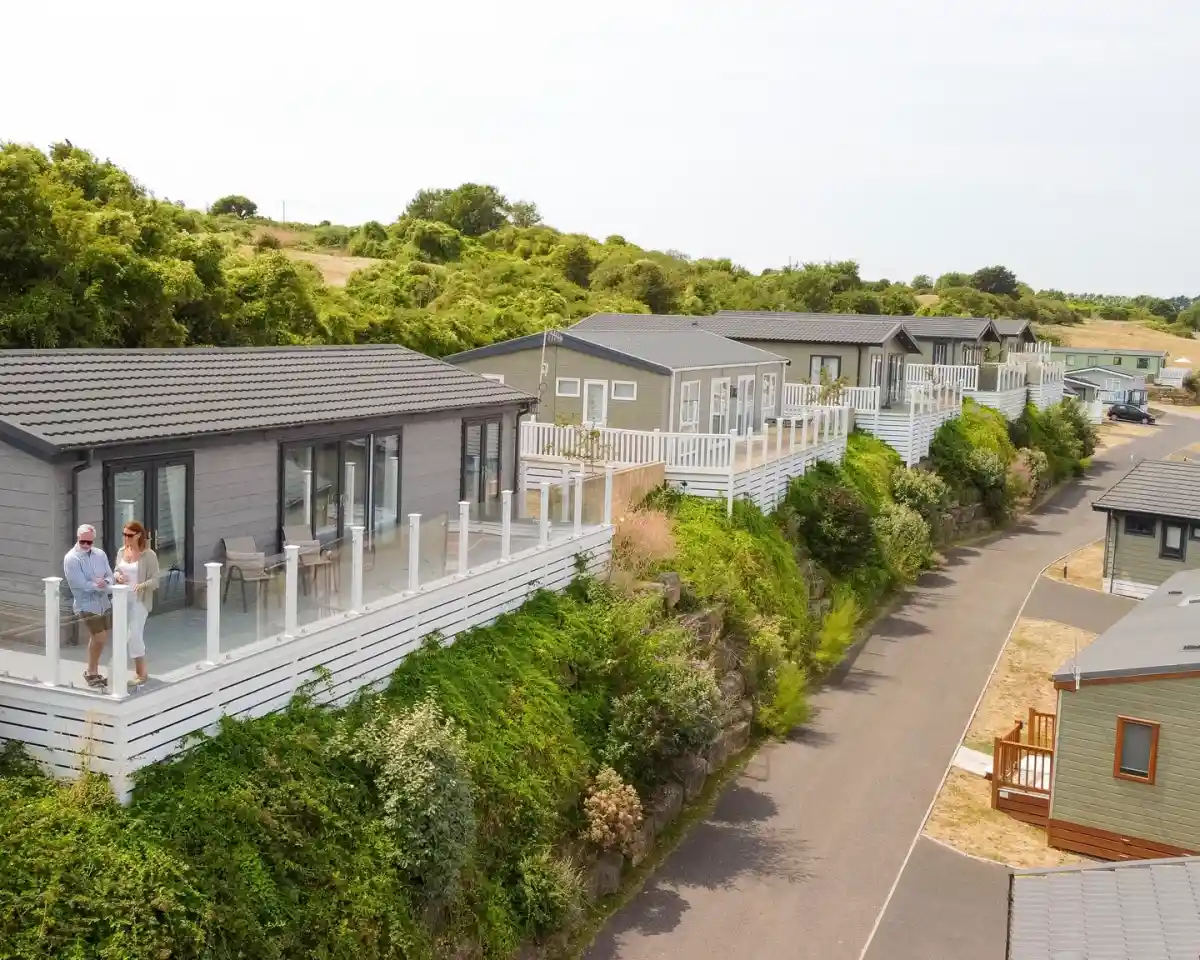 Aerial view of a row of modern holiday cabins with glass balconies, surrounded by greenery along a winding road. Two people stand on a balcony, enjoying the view.