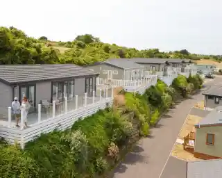 Aerial view of a row of modern holiday cabins with glass balconies, surrounded by greenery along a winding road. Two people stand on a balcony, enjoying the view.