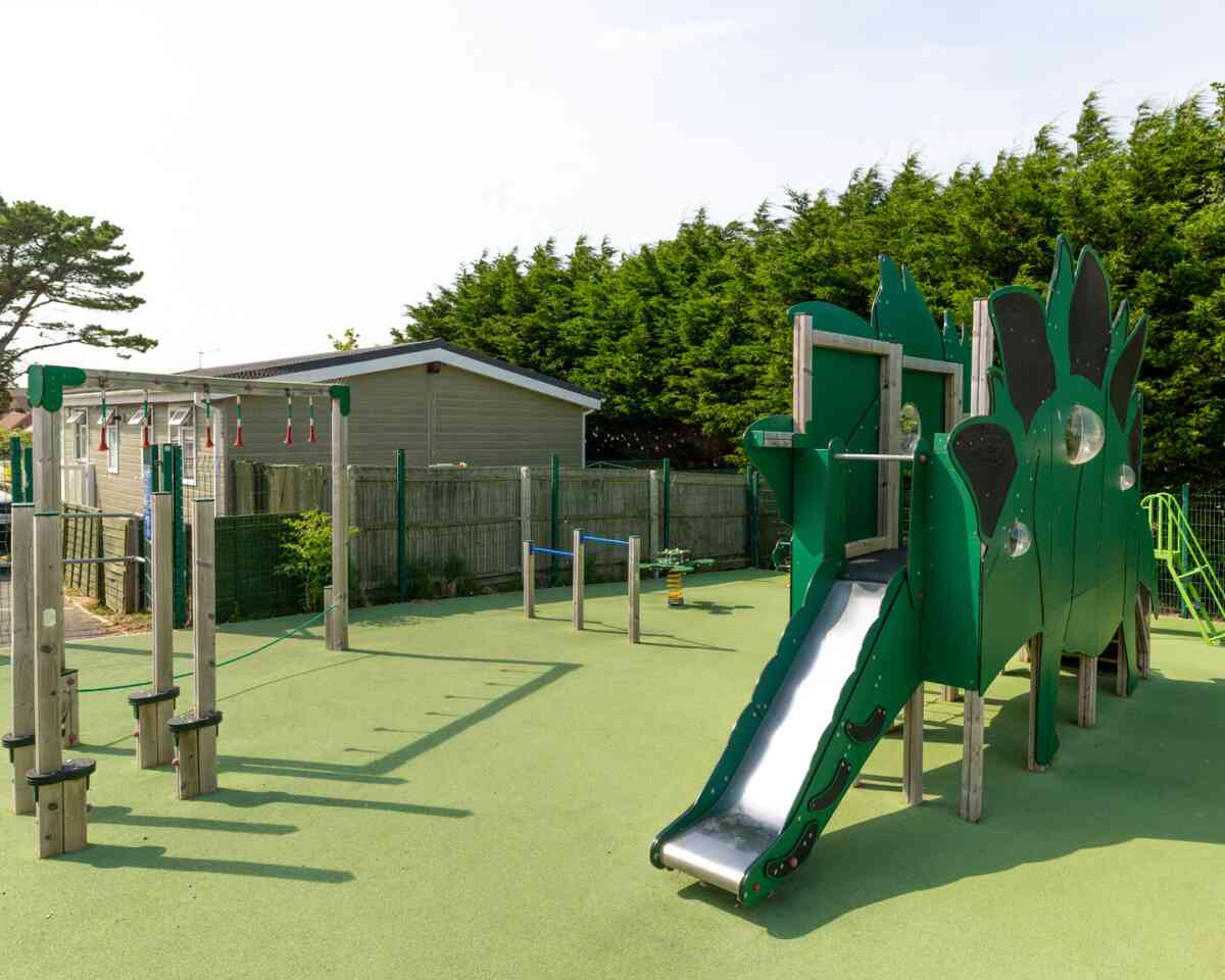 A colorful playground featuring a dragon-themed climbing structure with a slide, surrounded by green flooring and trees in the background. Various climbing and exercise apparatus are also visible.