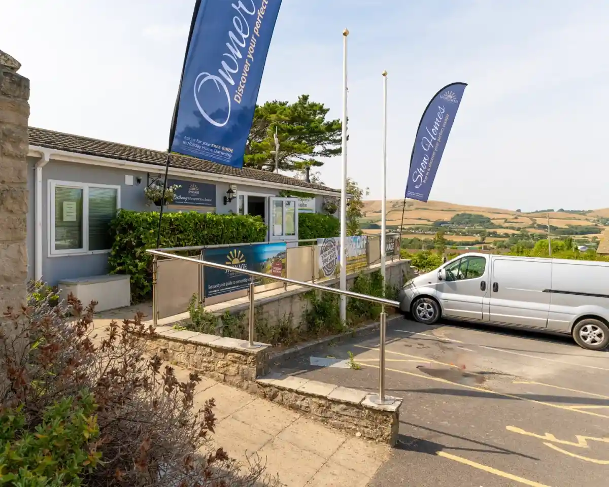 A building with a blue exterior featuring signs promoting ownership and discovery. Flags are waving in front, and a white van is parked nearby. Rolling hills are visible in the background under a clear sky.