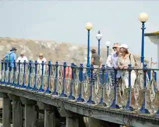 A couple leans against the railing of a pier, smiling at each other. In the background, various people walk along the pier, with a scenic view of a hillside and the coastline. White lamps line the pier, creating a charming atmosphere.