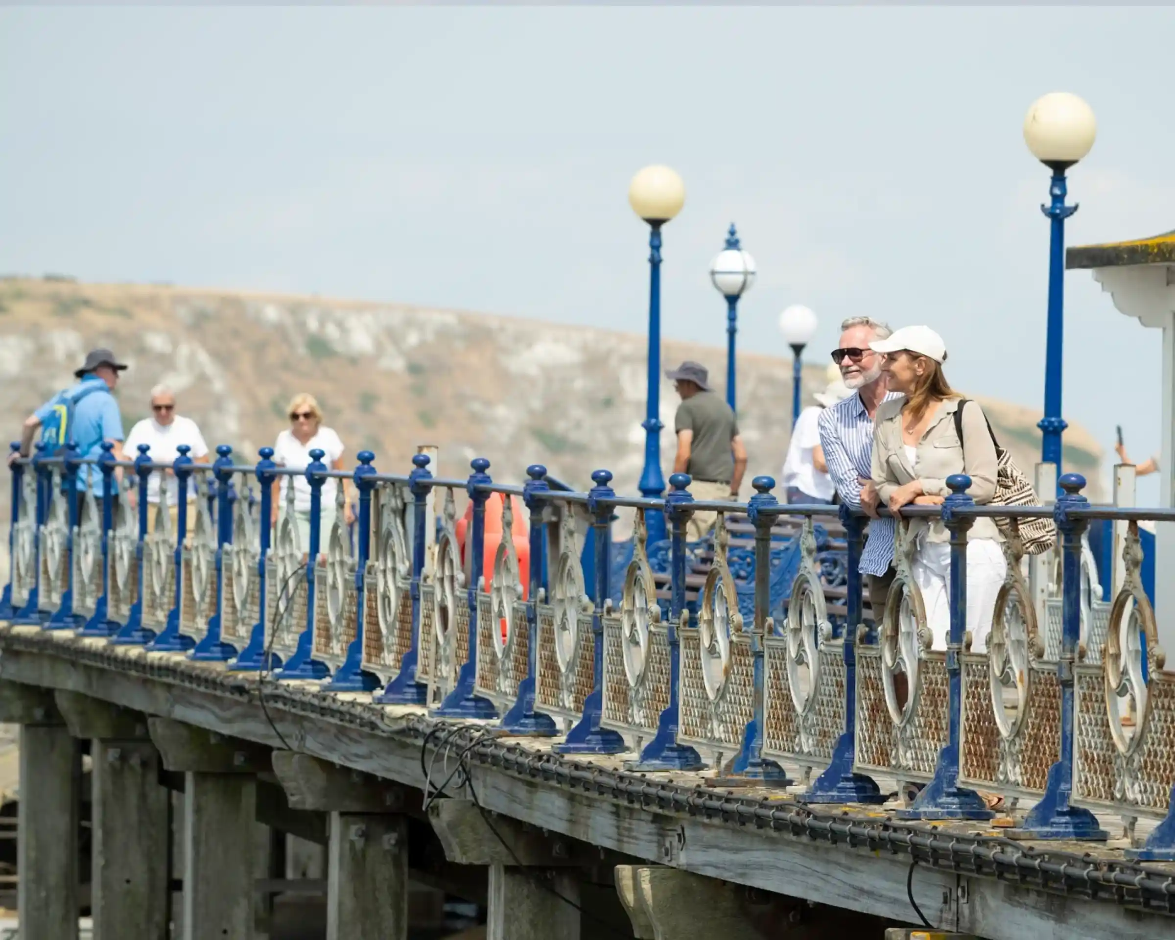 A couple leans against the railing of a pier, smiling at each other. In the background, various people walk along the pier, with a scenic view of a hillside and the coastline. White lamps line the pier, creating a charming atmosphere.