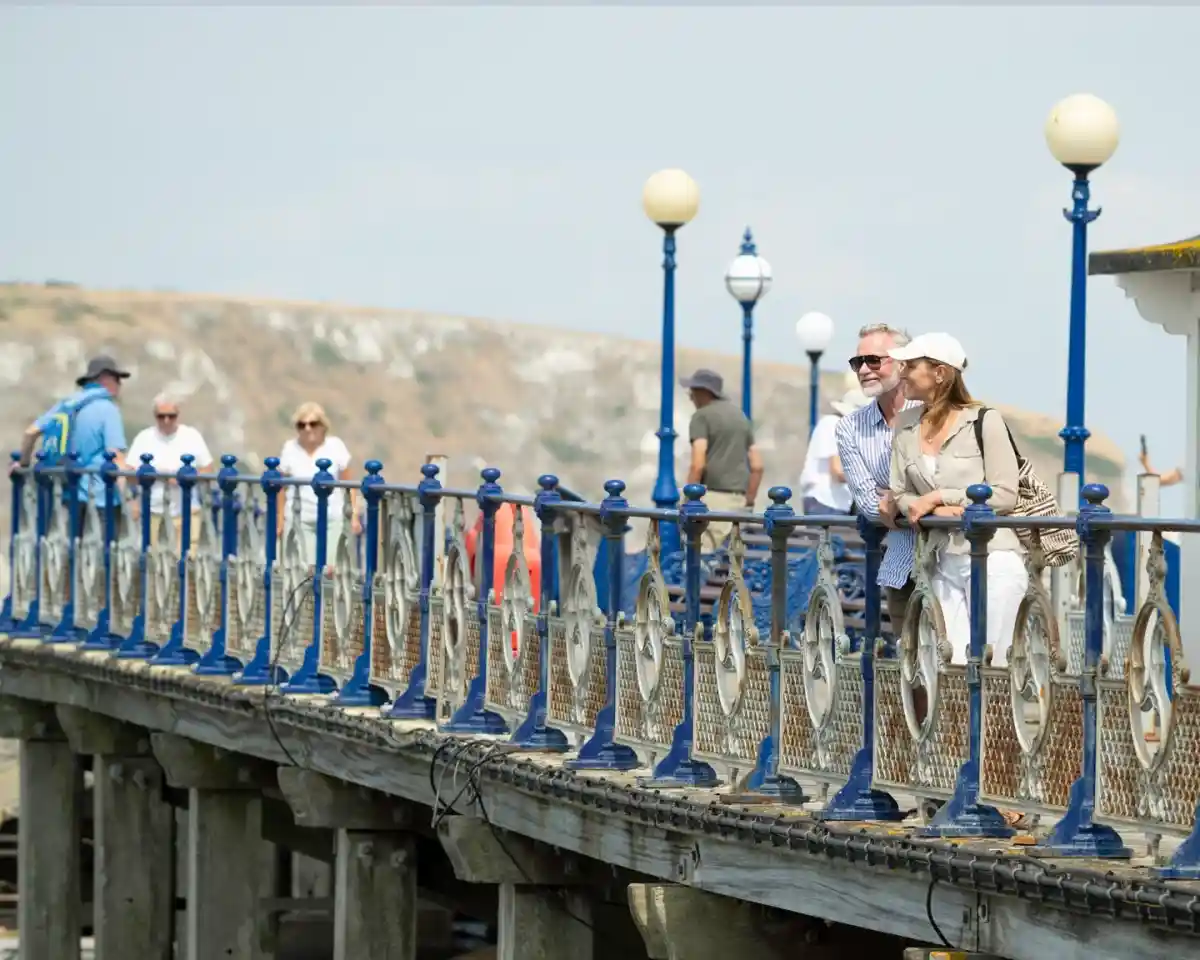 A couple leans against the railing of a pier, smiling at each other. In the background, various people walk along the pier, with a scenic view of a hillside and the coastline. White lamps line the pier, creating a charming atmosphere.