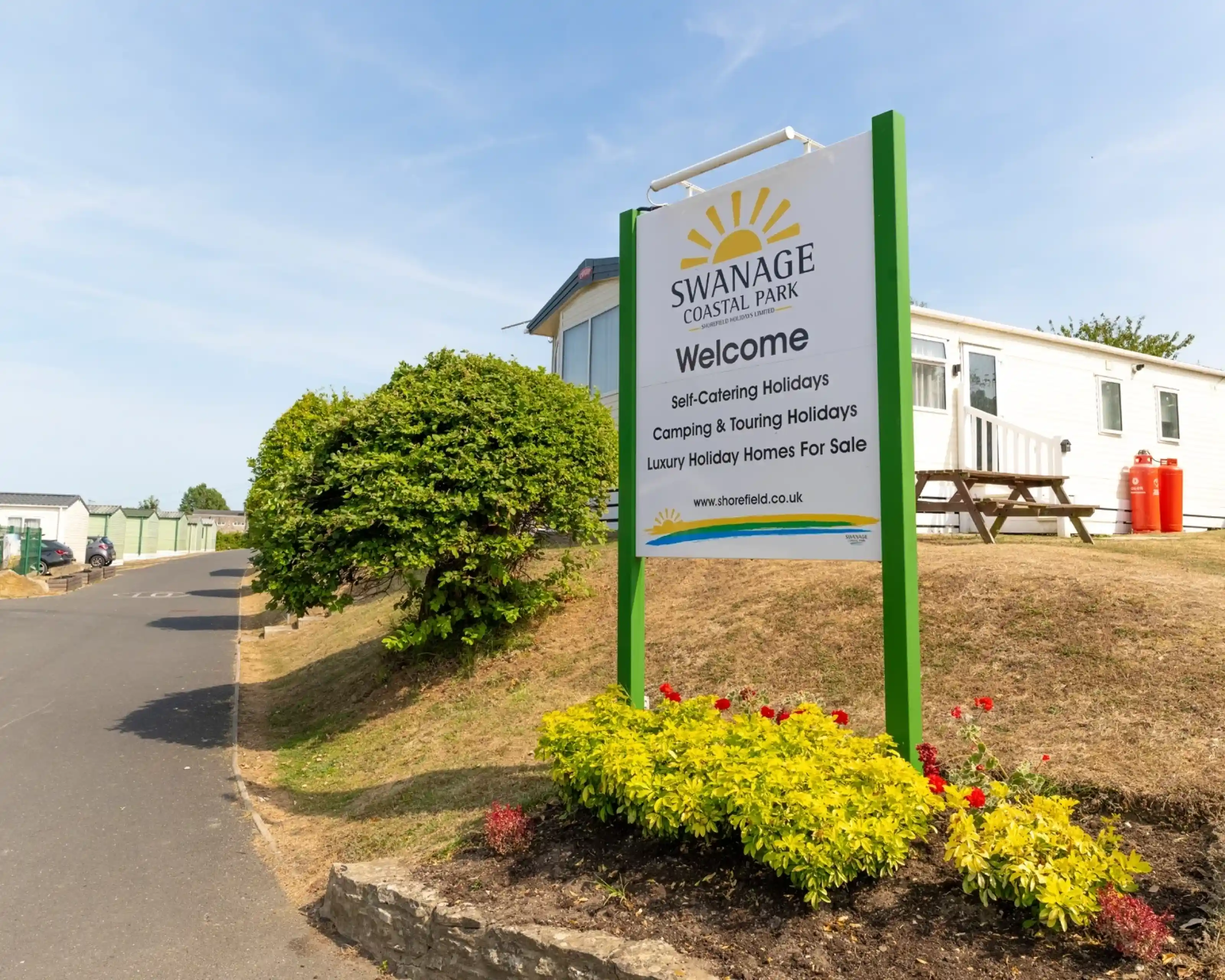 Sign for Swanage Coastal Park welcoming visitors, featuring details about self-catering holidays, camping, and luxury holiday homes for sale. The sign is surrounded by colorful flowers and greenery, with a view of holiday accommodations in the background.