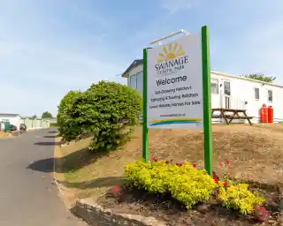 Sign for Swanage Coastal Park welcoming visitors, featuring details about self-catering holidays, camping, and luxury holiday homes for sale. The sign is surrounded by colorful flowers and greenery, with a view of holiday accommodations in the background.