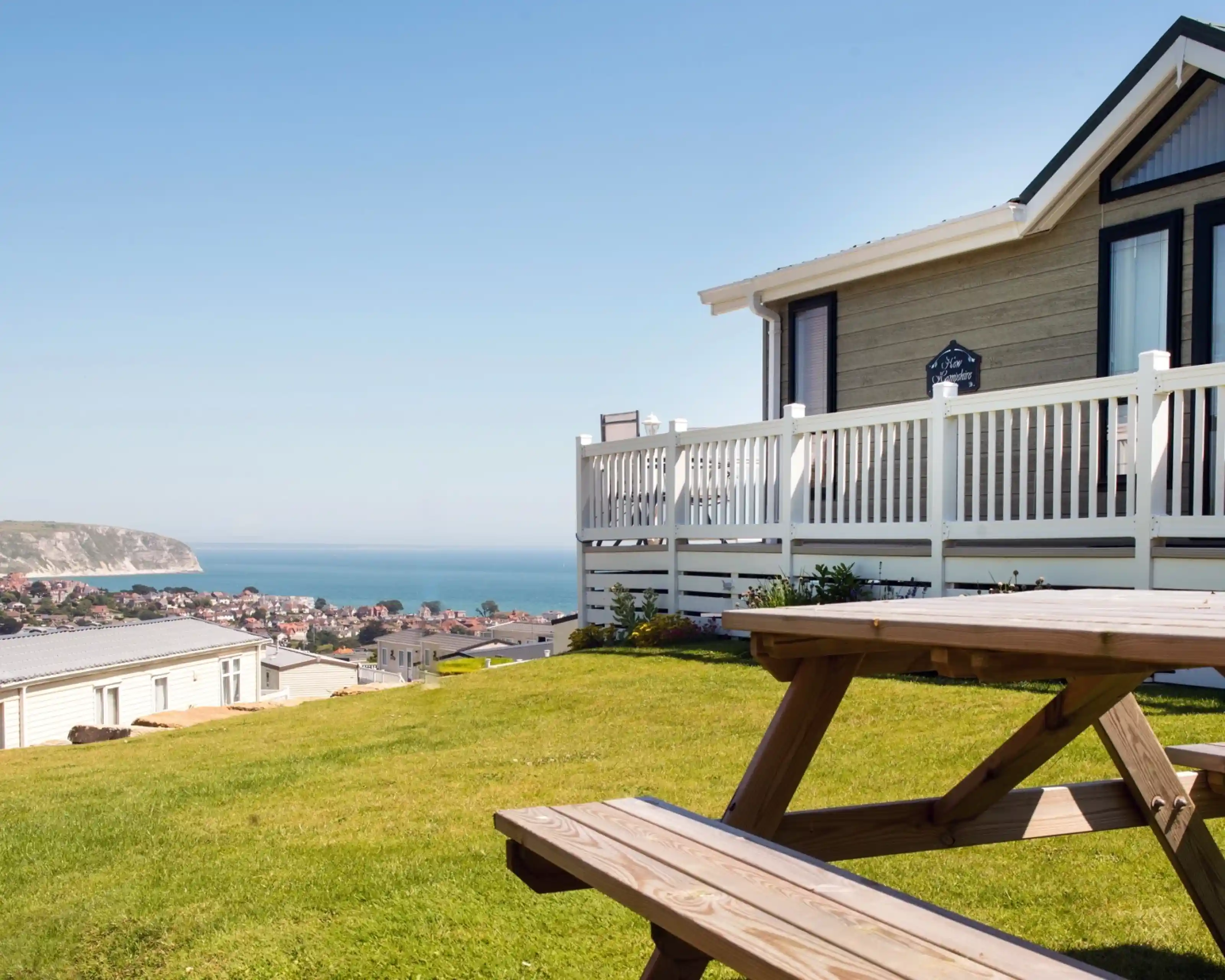 A coastal view from a property, featuring a wooden picnic table in the foreground and a white railing. The landscape includes lush green grass, a clear blue sky, and the ocean in the distance, with a small town visible along the shore.