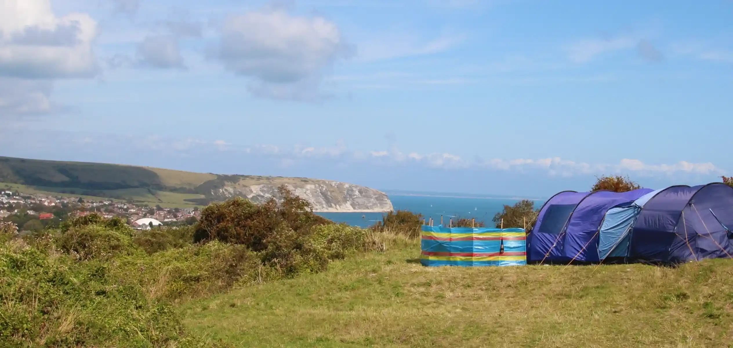 Tents set up in a grassy area with a view of a coastline and ocean in the background. Rolling hills and clouds fill the sky.