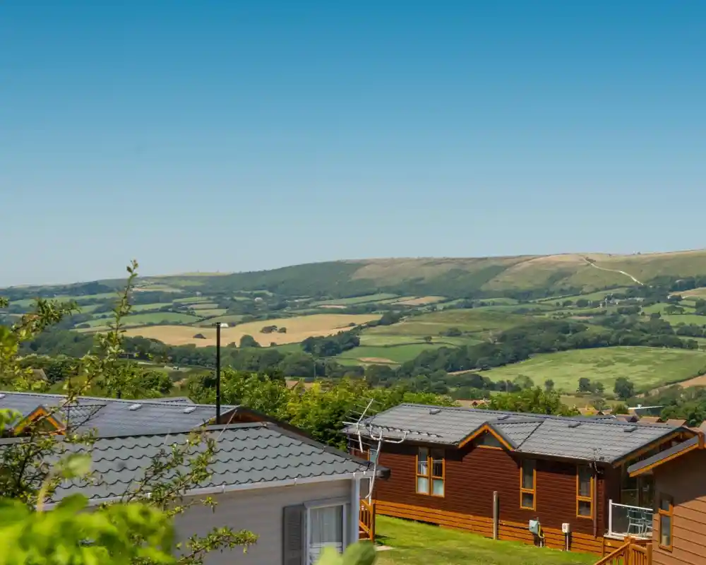 A scenic view of rolling green hills and farmland under a clear blue sky, with two wooden lodges in the foreground.