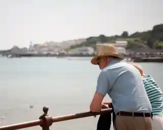 An elderly man in a straw hat leans over a railing, gazing out at the water. A woman beside him, wearing a striped shirt, also looks at the serene scene. In the background, a coastal town with white buildings can be seen along the shoreline.