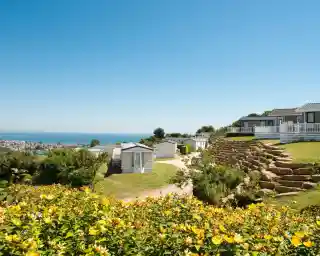 A scenic view of a coastal campsite featuring colorful flowers in the foreground, neatly arranged caravans on a hillside, and the ocean visible in the background under a clear blue sky.
