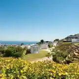 A scenic view of a coastal campsite featuring colorful flowers in the foreground, neatly arranged caravans on a hillside, and the ocean visible in the background under a clear blue sky.