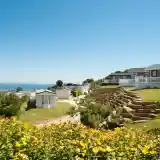A scenic view of a coastal campsite featuring colorful flowers in the foreground, neatly arranged caravans on a hillside, and the ocean visible in the background under a clear blue sky.