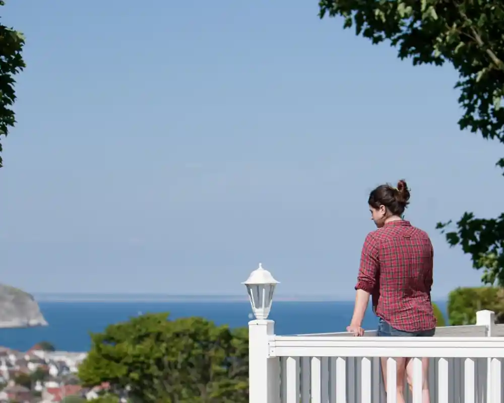 A woman in a red plaid shirt stands on a white balcony railing, looking out toward the ocean. Lush greenery surrounds her, and the coastline is visible in the background under a clear blue sky.