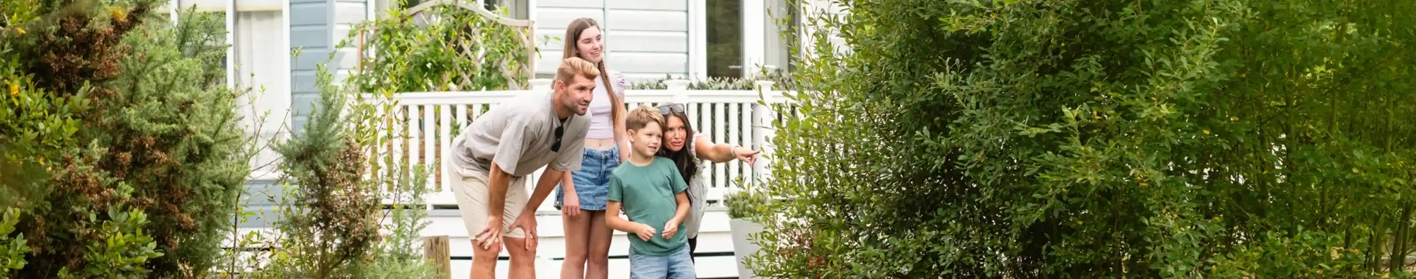 A family of four, including two adults and two children, leans over a wooden dock by a pond covered with lily pads, looking at something in the water. A blue lodge is in the background, surrounded by greenery.