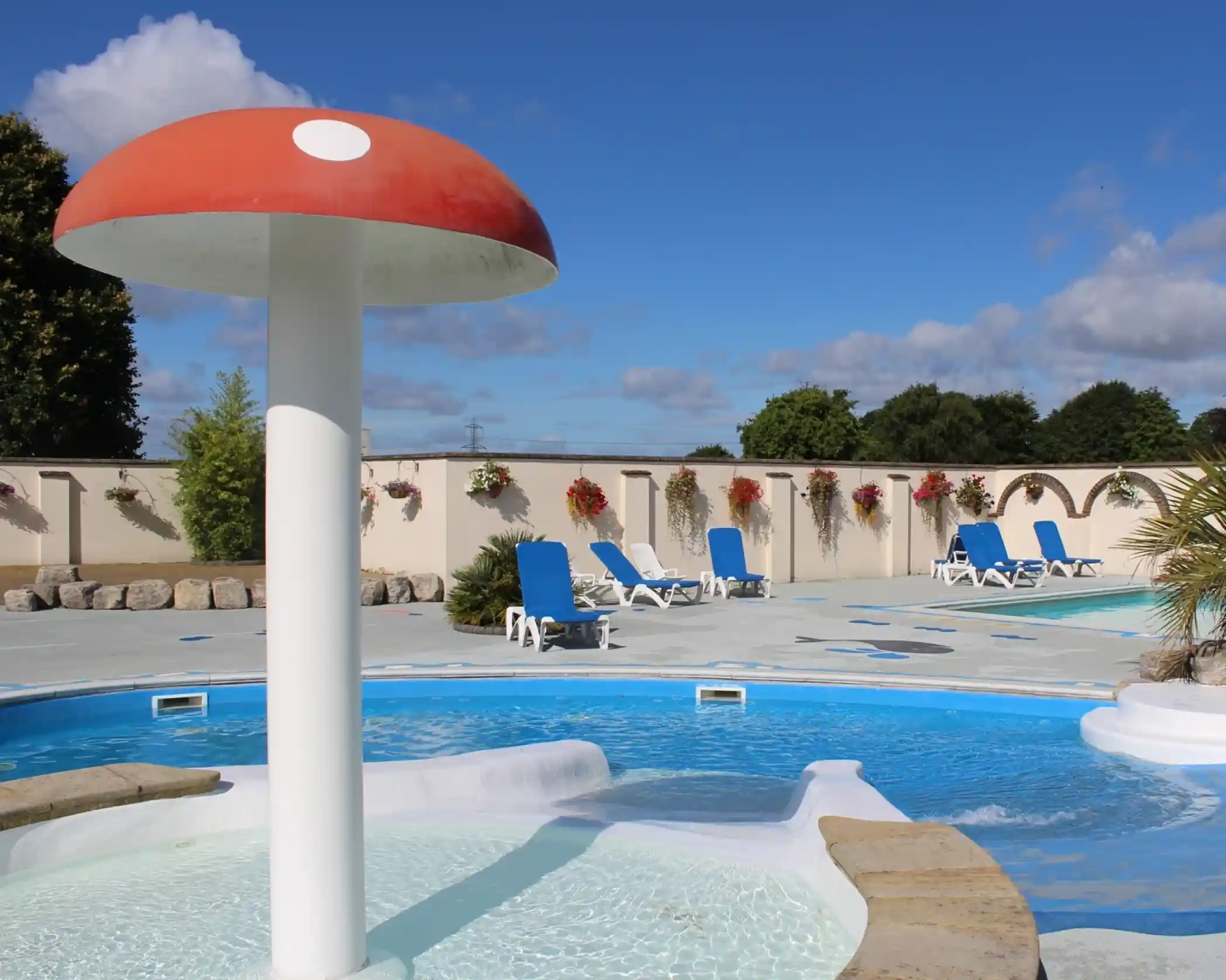 A scenic outdoor pool area featuring a mushroom-shaped water feature surrounded by lounge chairs and decorative flowers against a backdrop of blue skies and fluffy clouds.