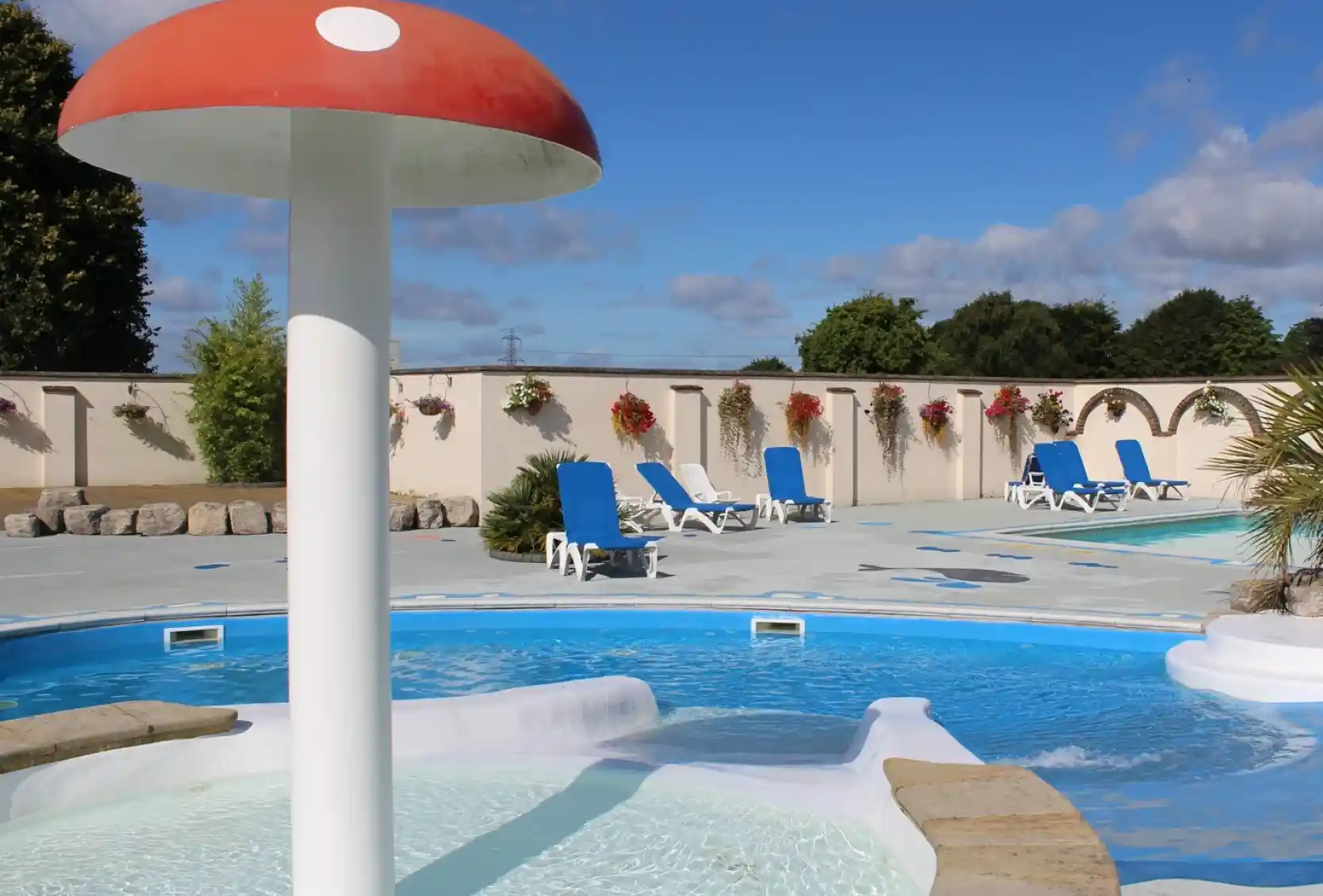 A scenic outdoor pool area featuring a mushroom-shaped water feature surrounded by lounge chairs and decorative flowers against a backdrop of blue skies and fluffy clouds.