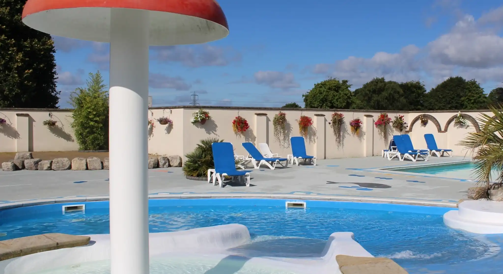 A scenic outdoor pool area featuring a mushroom-shaped water feature surrounded by lounge chairs and decorative flowers against a backdrop of blue skies and fluffy clouds.