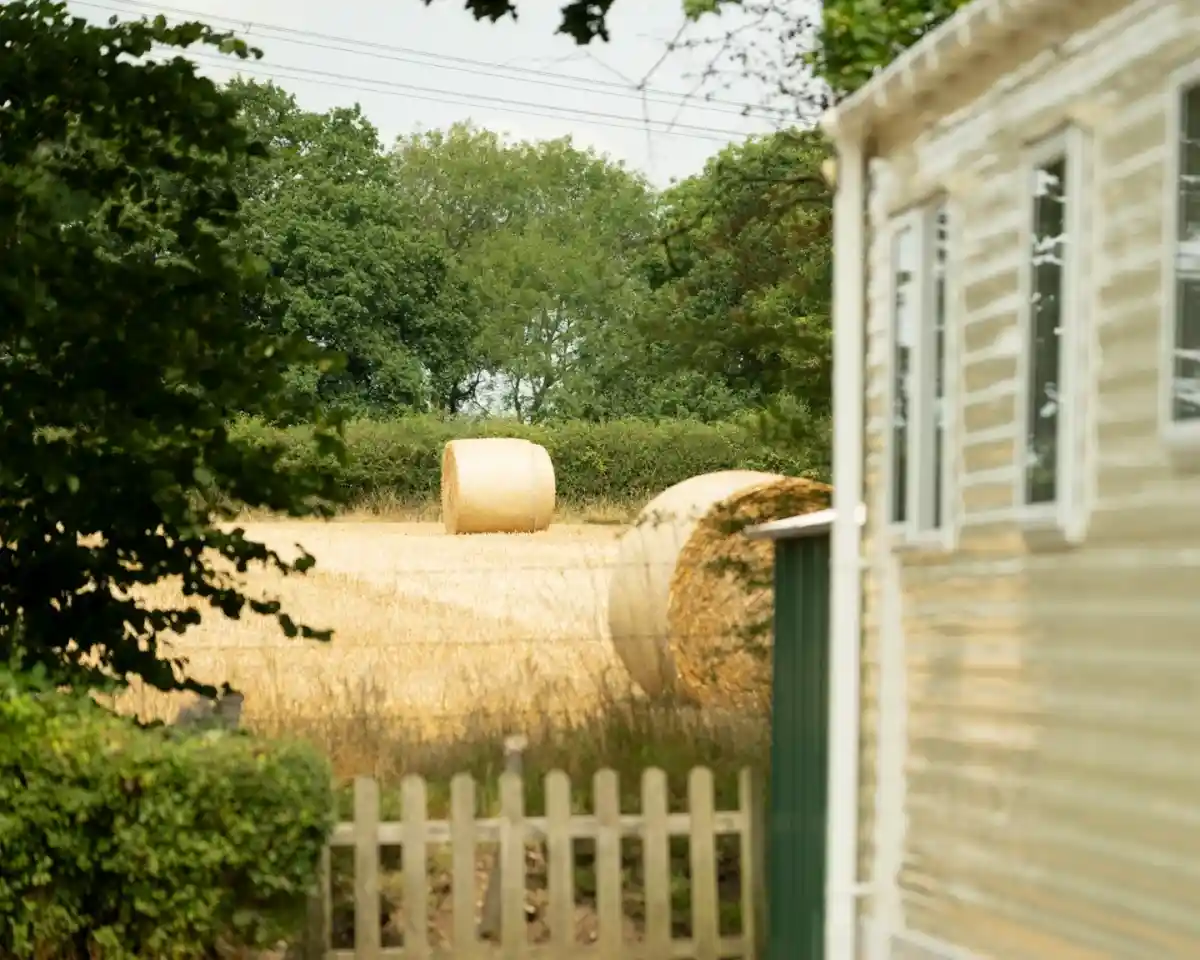 A golden hayfield with two round hay bales in the background, framed by trees and a small fence. A mobile home is partially visible to the right.