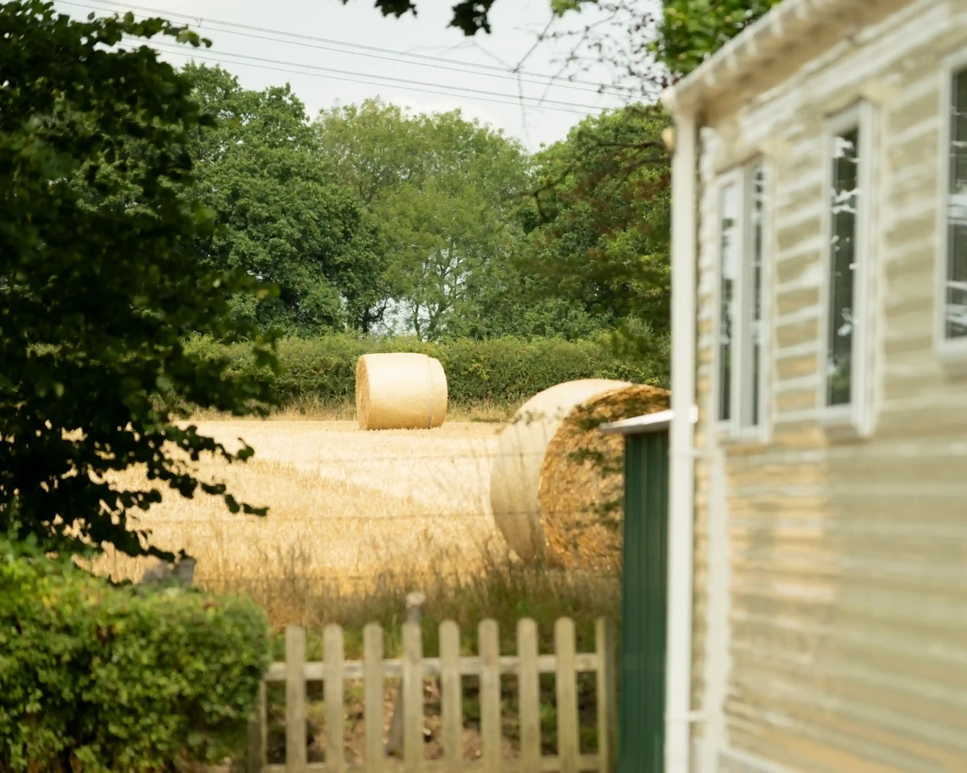 A golden hayfield with two round hay bales in the background, framed by trees and a small fence. A mobile home is partially visible to the right.