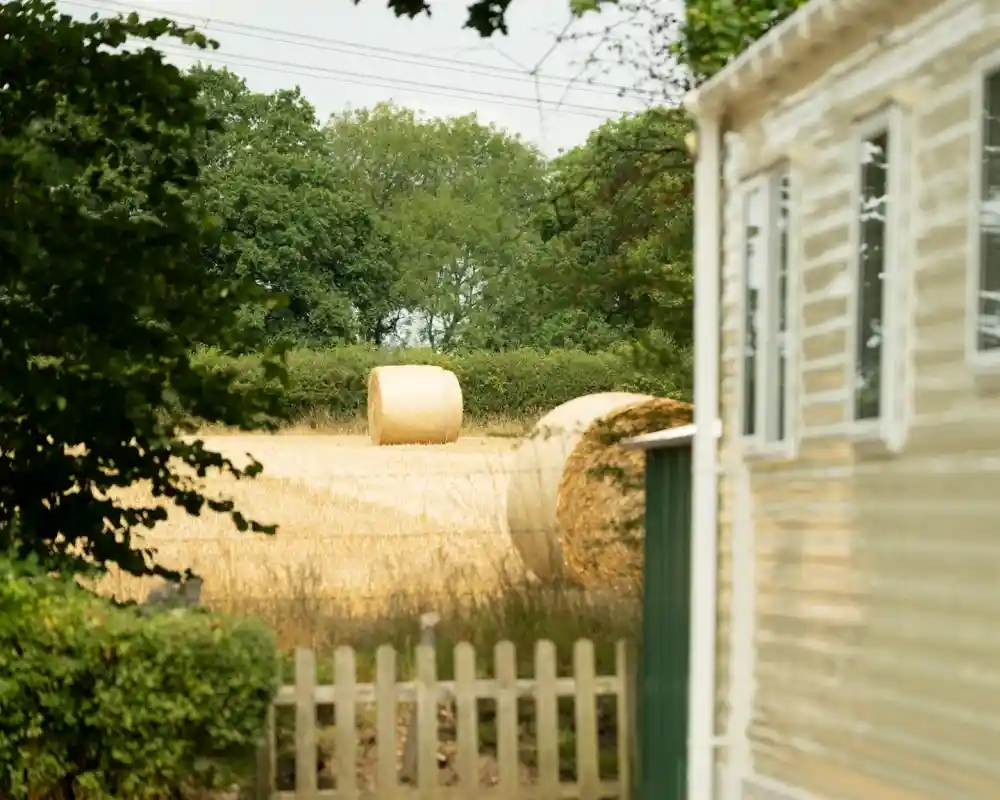 A golden hayfield with two round hay bales in the background, framed by trees and a small fence. A mobile home is partially visible to the right.